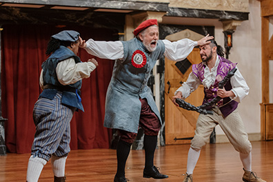 ASC-Comedy_Errors-Dromio and Antipholus of Ephesus Dromio of Ephesus in boxing pose wearing blue vest and hat, blue striped kneepants with with stockings and work boots faces off against Antopholus of Ephesus holding a short black rope in his hands and wearing a fancy purple vest over white shirt and cream kneepants with white stockings and stylish shoes. The officer stands between them with outstretched hands planted on their foreheads; he's wearing a long vest coat with a red ribboned emplem, red cap, and maroon knee pants with black stockings.